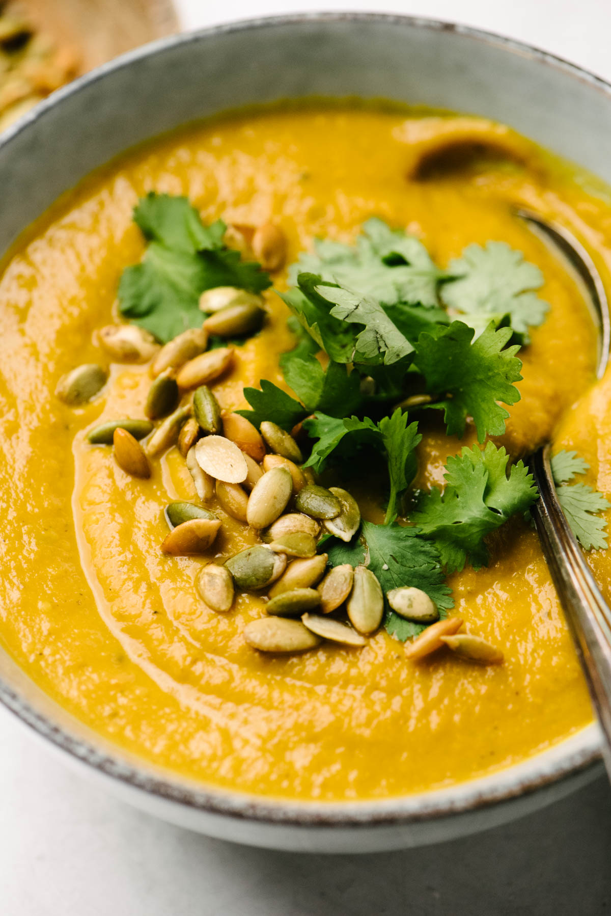 Side view, a soup spoon tucked into a bowl of pumpkin soup with curry, garnished with pepitas and fresh cilantro.