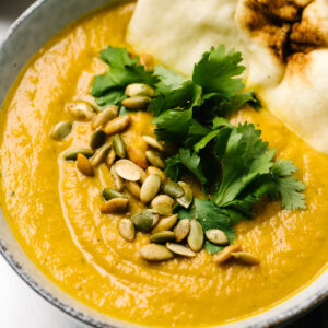 Side view, a piece of naan dipped into a bowl of pumpkin curry soup, garnished with pepitas and cilantro.