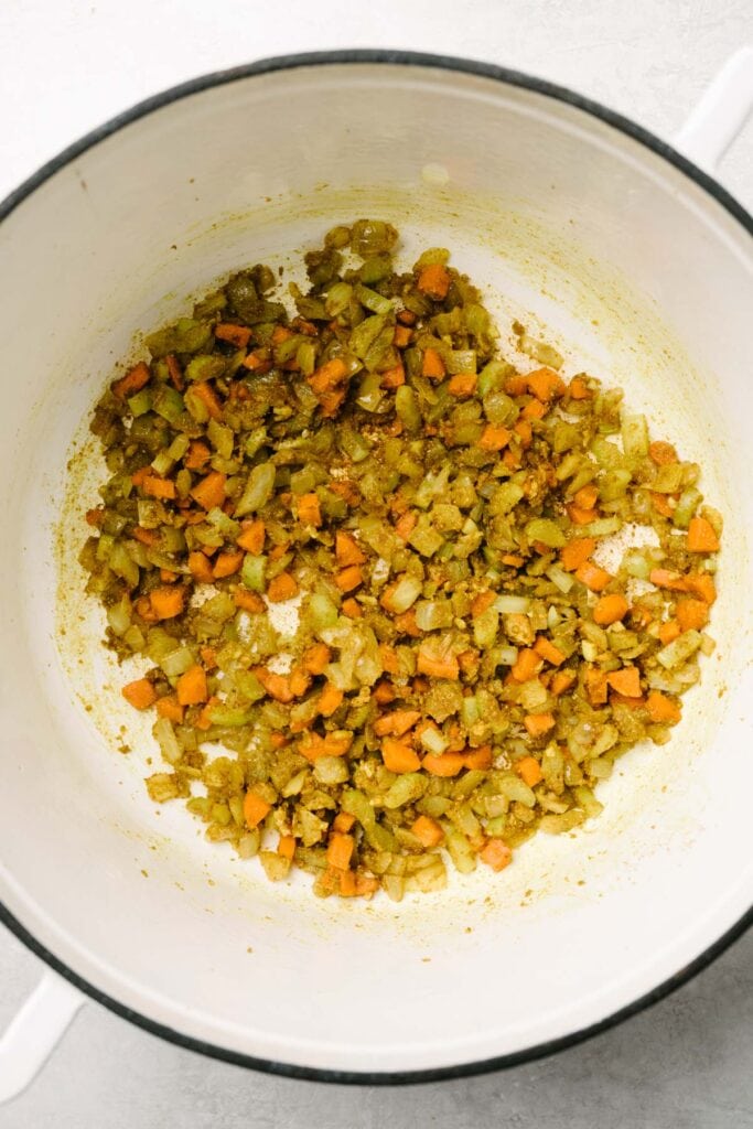 Diced carrots, onions, and celery with curry spices in a white dutch oven.