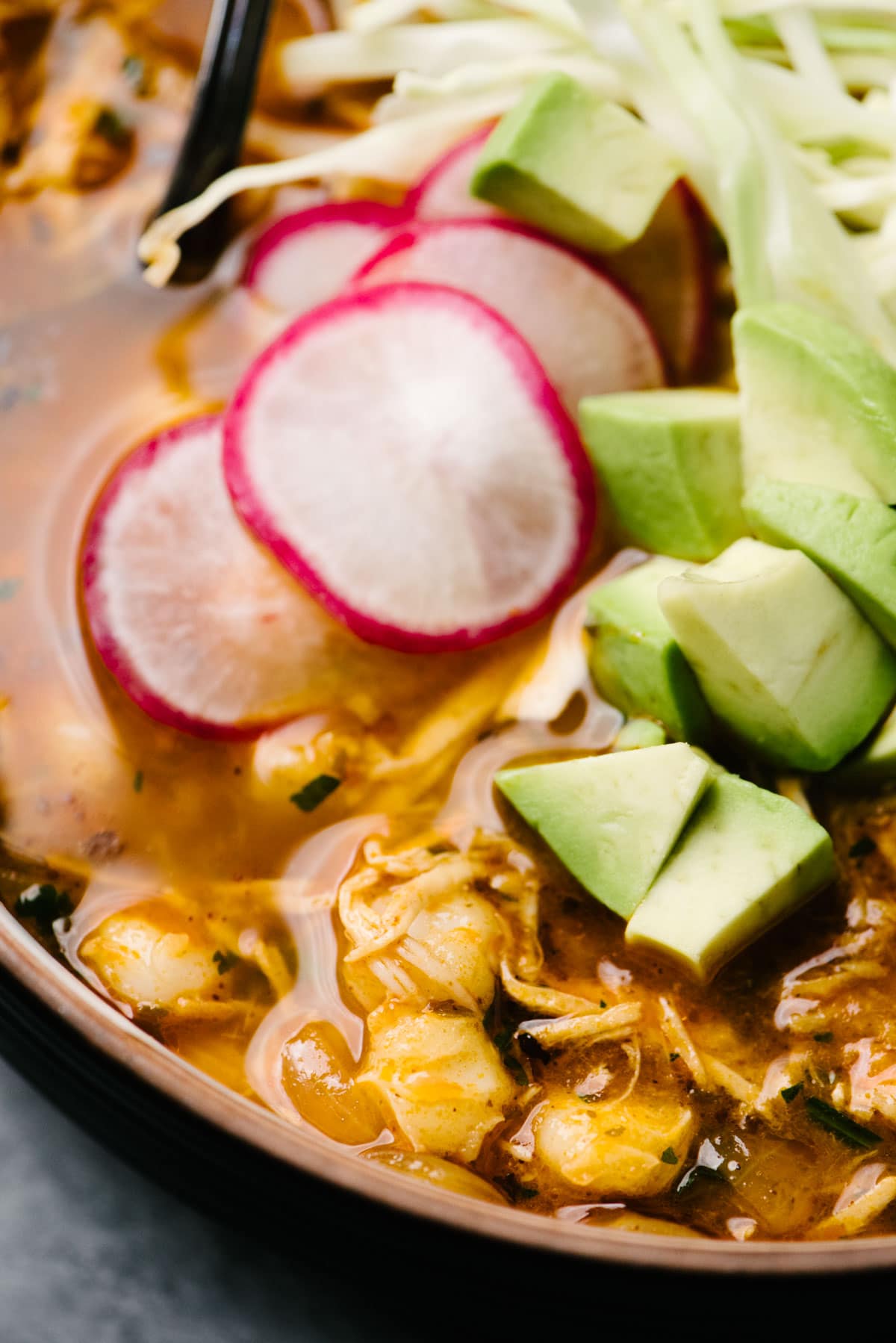 Close up side view of a bowl of turkey pozole garnished with radish slices, avocado, cabbage, and lime wedges.
