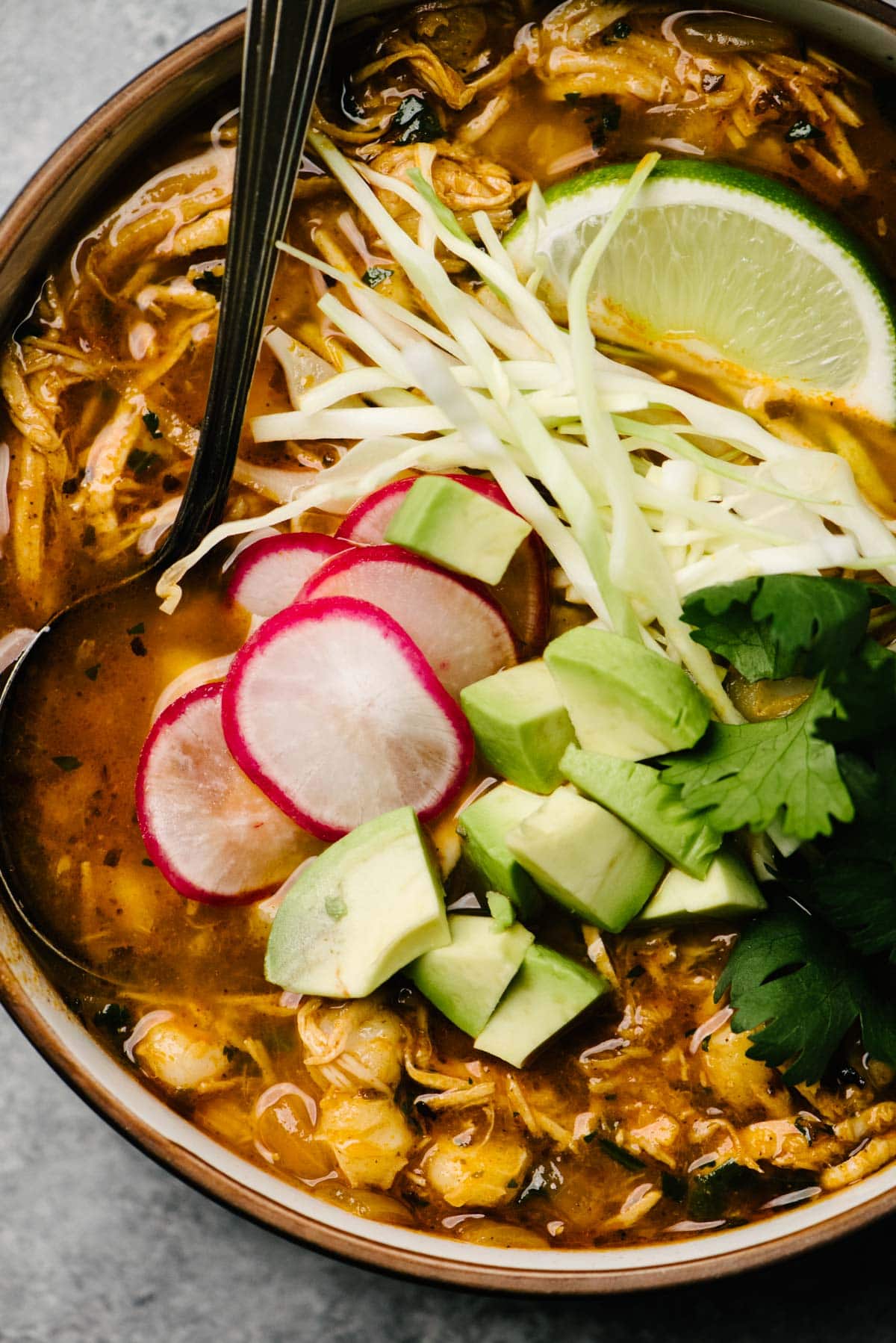 A spoon tucked into a bowl of turkey pozole topped with cabbage, cilantro, avocado, lime wedges, and radish slices.