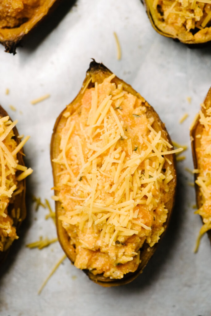 Sweet potato halves on a a baking sheet, re-filed with the flesh and other ingredients, topped with shredded cheese. 