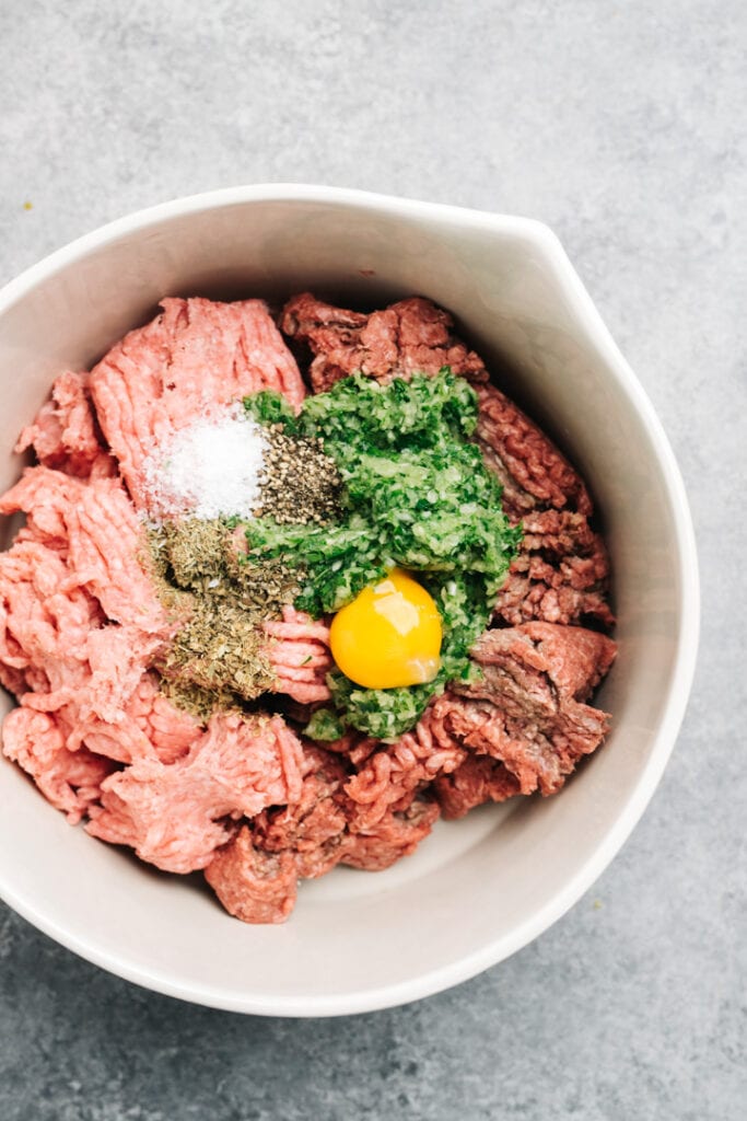 Ground beef, ground pork, egg yolk, fresh herbs, and dried spices in a large mixing bowl.