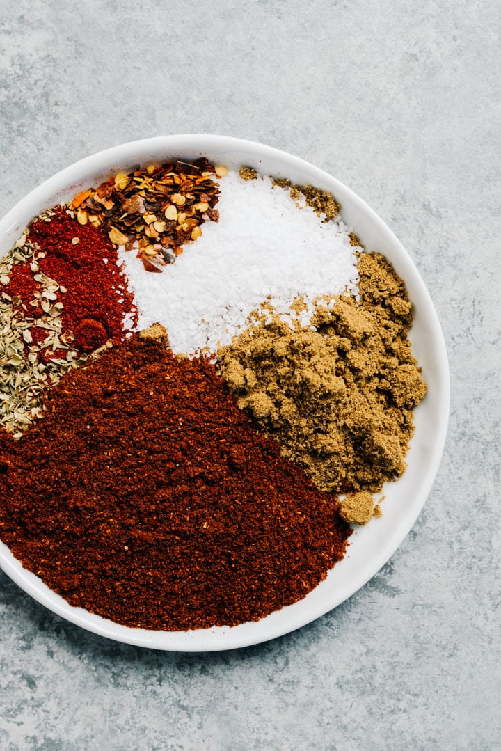 Chili seasonings on a small white plate - chili powder, cumin, oregano, paprika, kosher salt, and red pepper flakes..