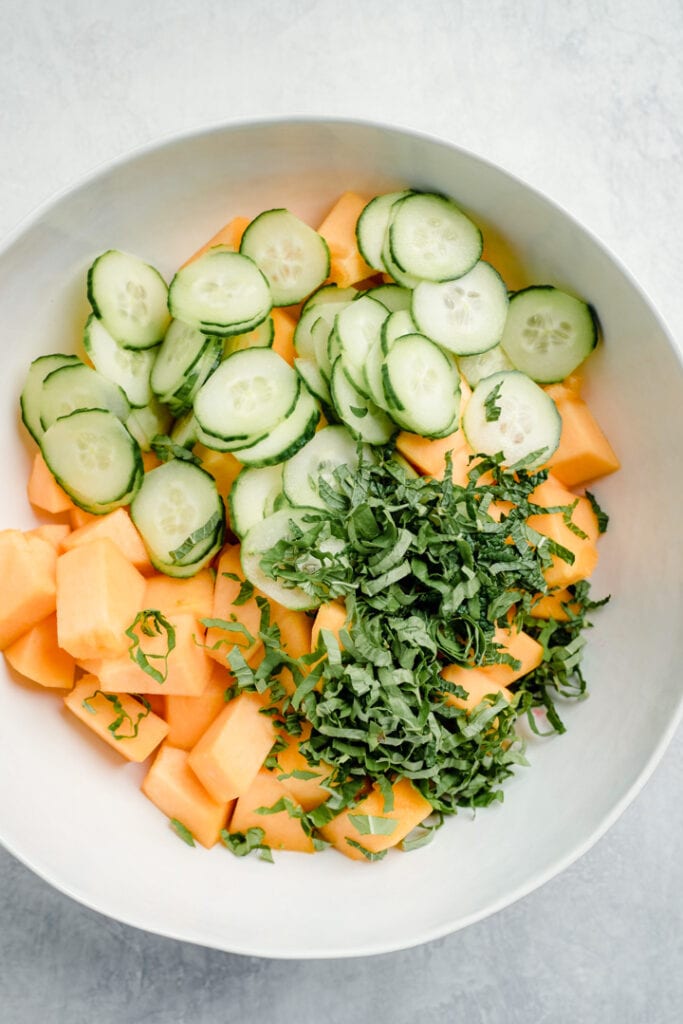 Diced cantaloupe, sliced cucumbers, and thinly sliced fresh mint and basil in a large white mixing bowl.