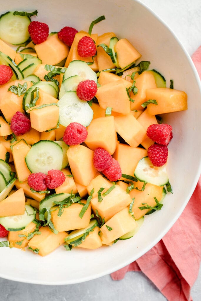 A cantaloupe salad with cucumbers, mint, basil, and raspberries in a large white bowl with a salmon colored napkin to the side.