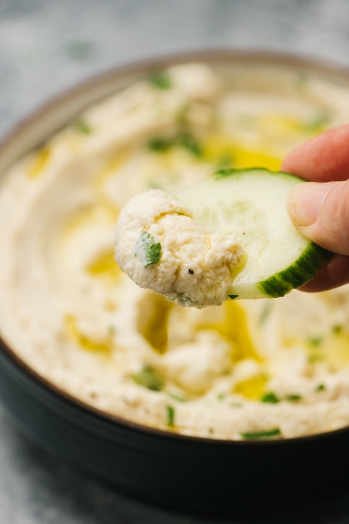 A hand holding a slice of cucumber dipped into cauliflower hummus, hovering over a bowl of dip.