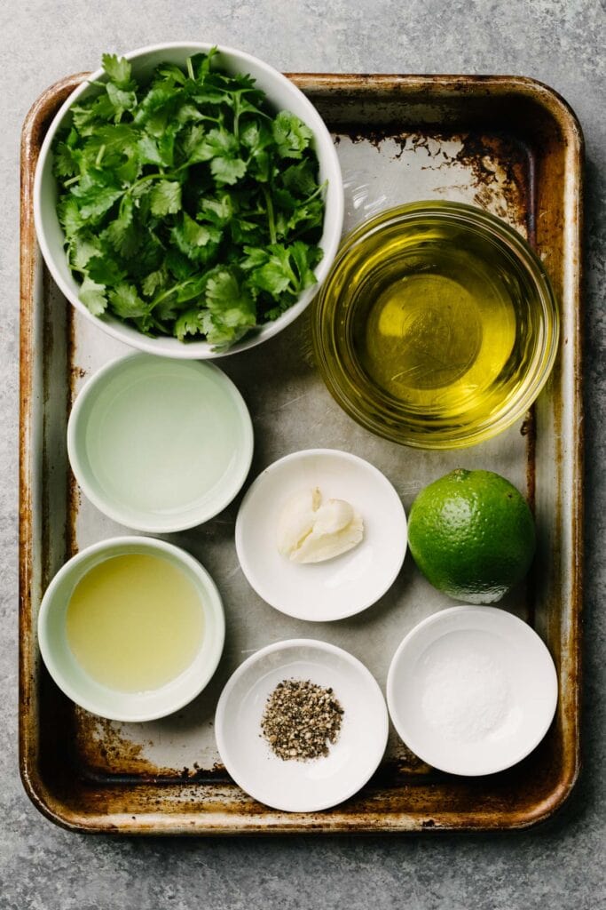 The ingredients for cilantro lime vinaigrette arranged in small bowls on a baking sheet - cilantro, olive oil, lime juice, white wine vinegar, garlic, salt, pepper, and honey.