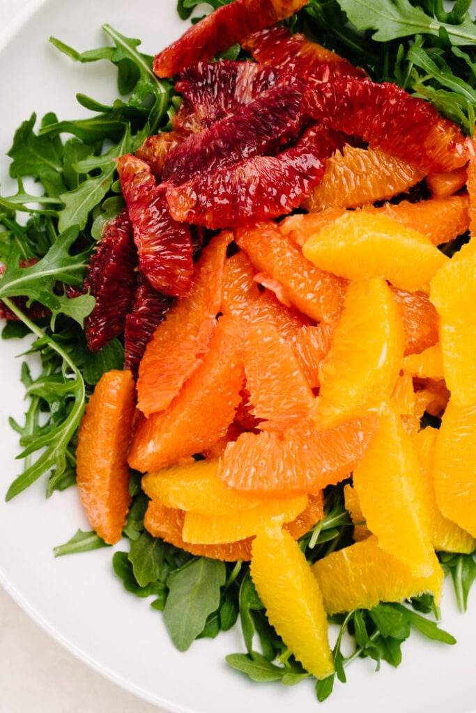 Arugula leaves topped with citrus fruits in a large white mixing bowl.