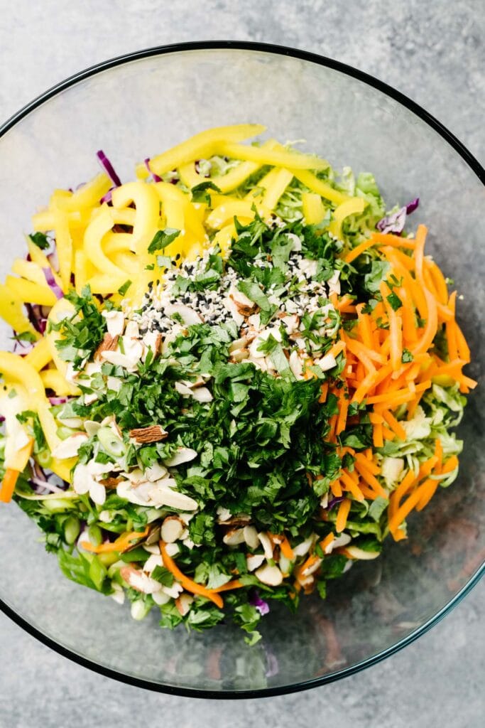 Shredded cabbage, shaved brussels sprouts, sliced bell pepper, shredded carrots, sesame seeds, almonds, and cilantro in a large glass mixing bowl.