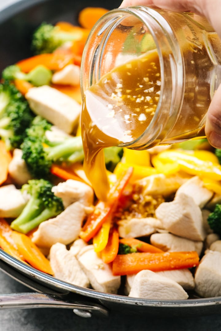 Pouring stir fry sauce over chicken and stir fry veggies in a skillet.