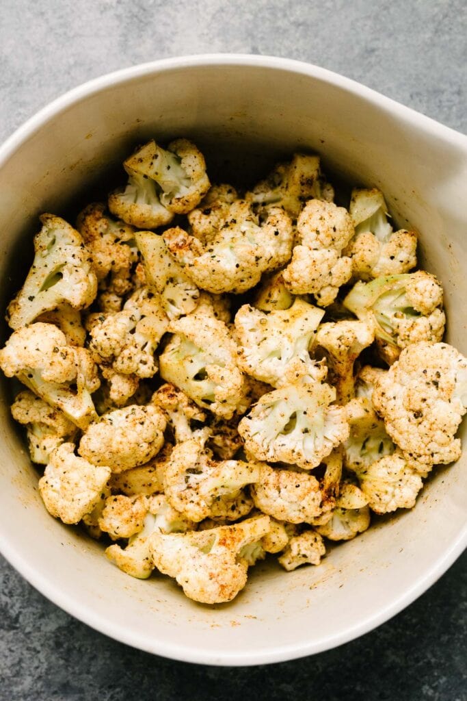 Cauliflower florets in a large mixing bowl tossed with olive oil, salt, and dried herbs and spices.