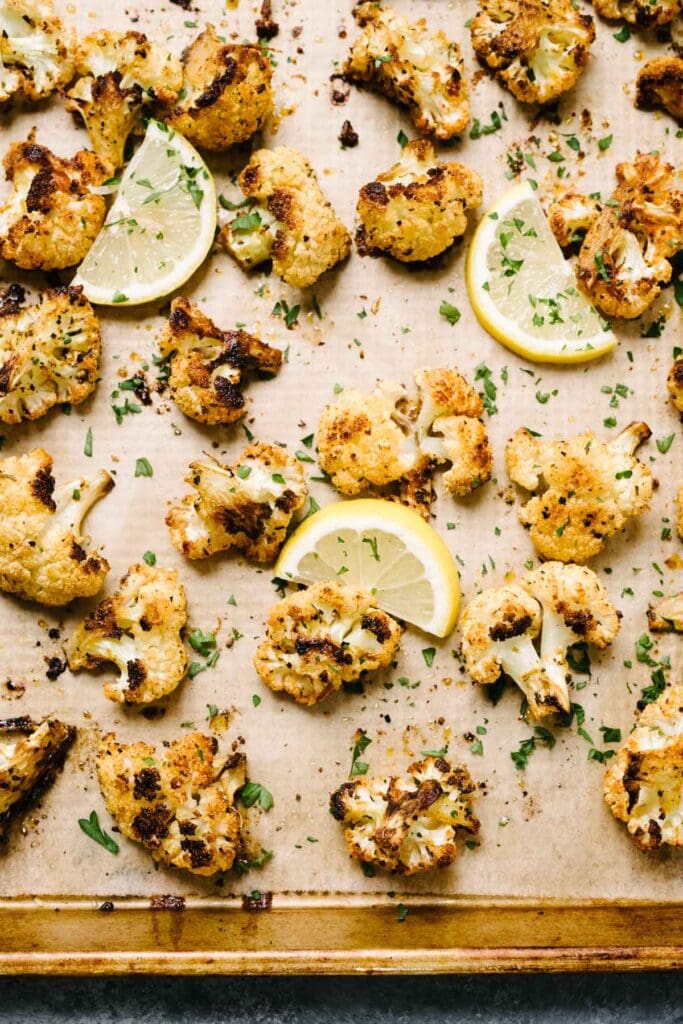 Roasted cauliflower florets on a parchment lined baking sheet, garnished with lemon wedges and fresh chopped parsley.