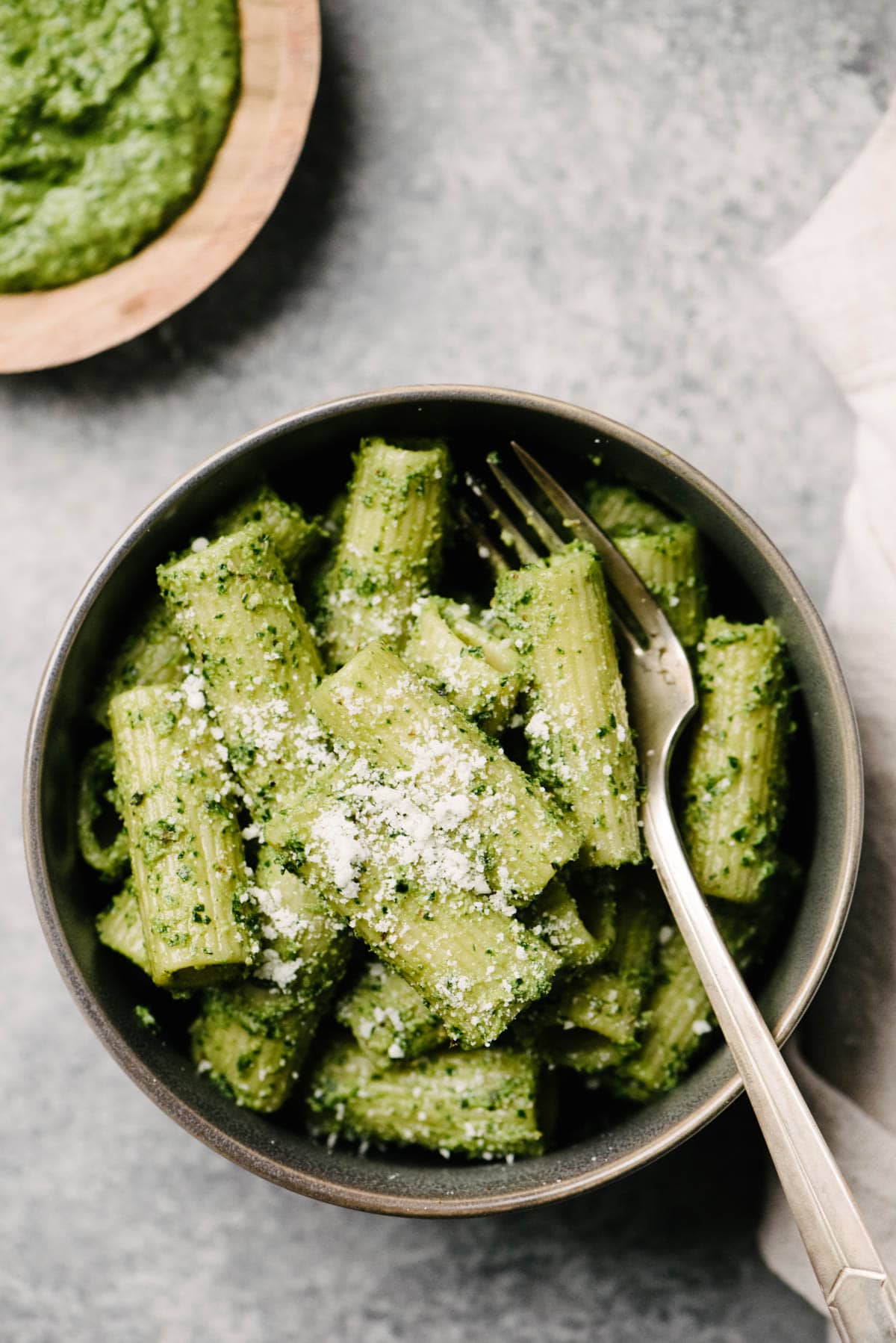 A bowl of rigatoni pasta tossed with spinach pesto on a concrete background with a linen napkin and small bowl of pesto to the side.