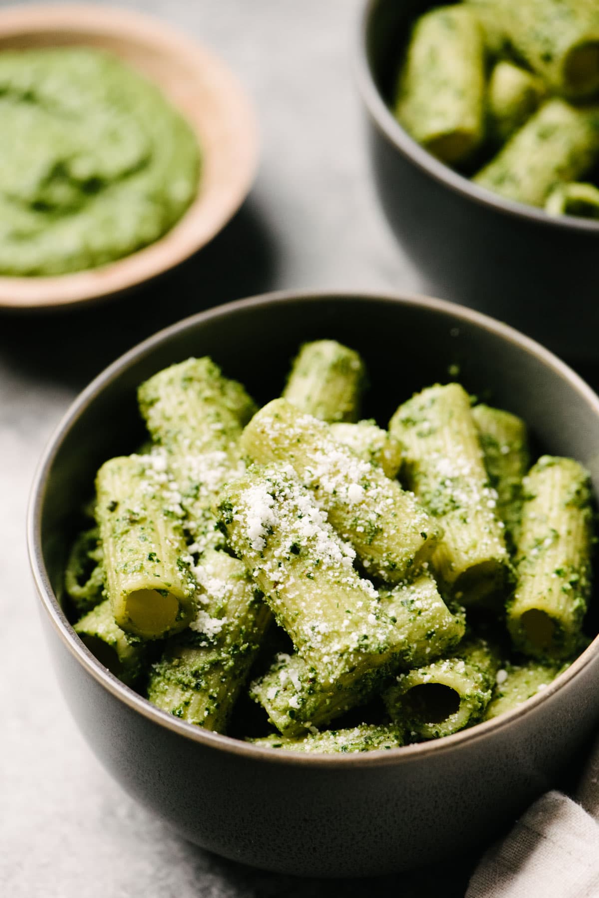 Side view, rigatoni tossed with creamy spinach pesto in grey bowls, with a small bowl of pesto in the background.