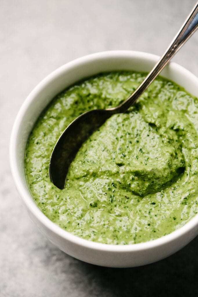 Side view, a spoon tucked into a small white bowl filled with creamy spinach pesto sauce.