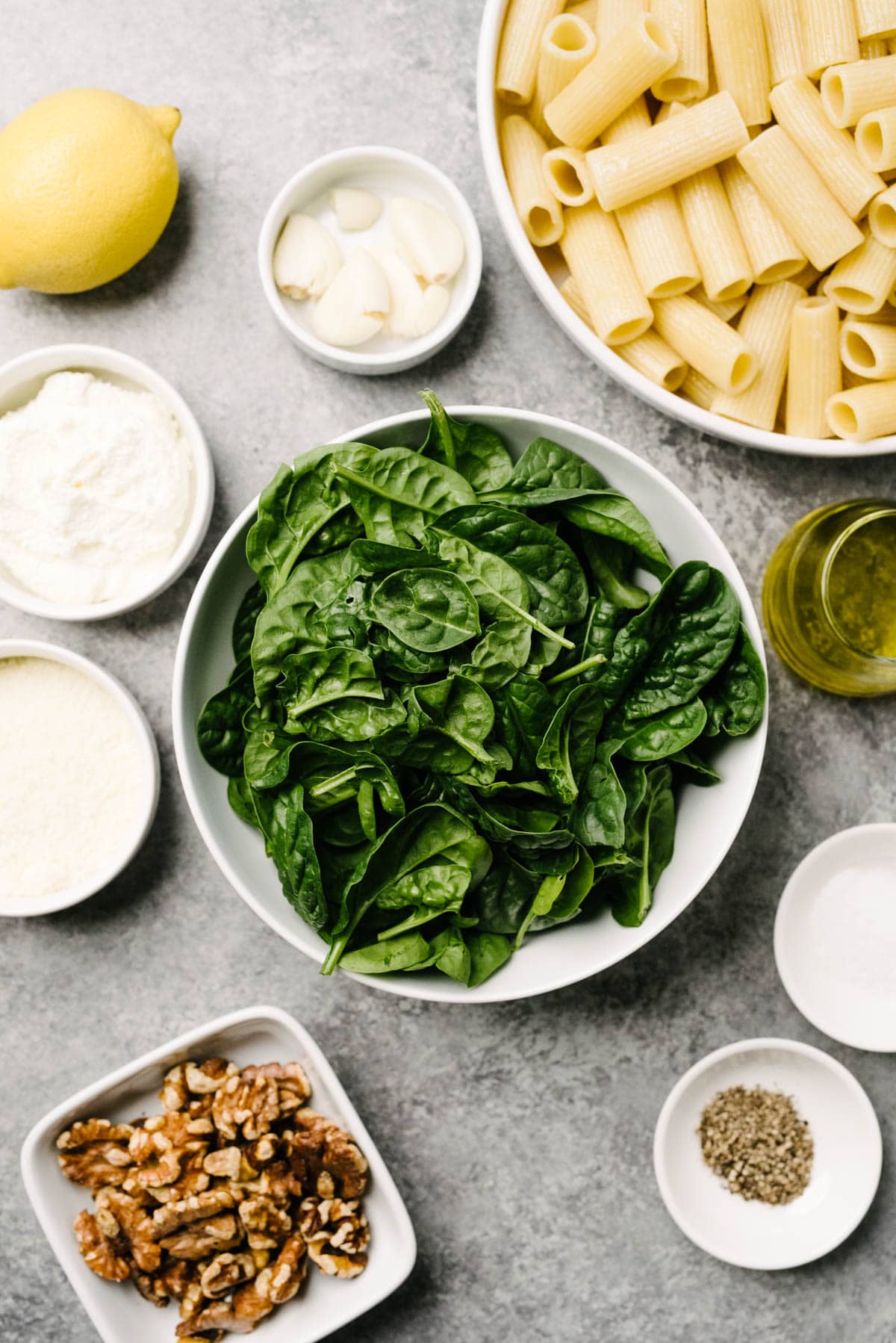The ingredients for spinach pesto on a concrete background - baby spinach, walnuts, ricotta, parmesan, lemon, garlic, salt, pepper, olive oil and cooked pasta.