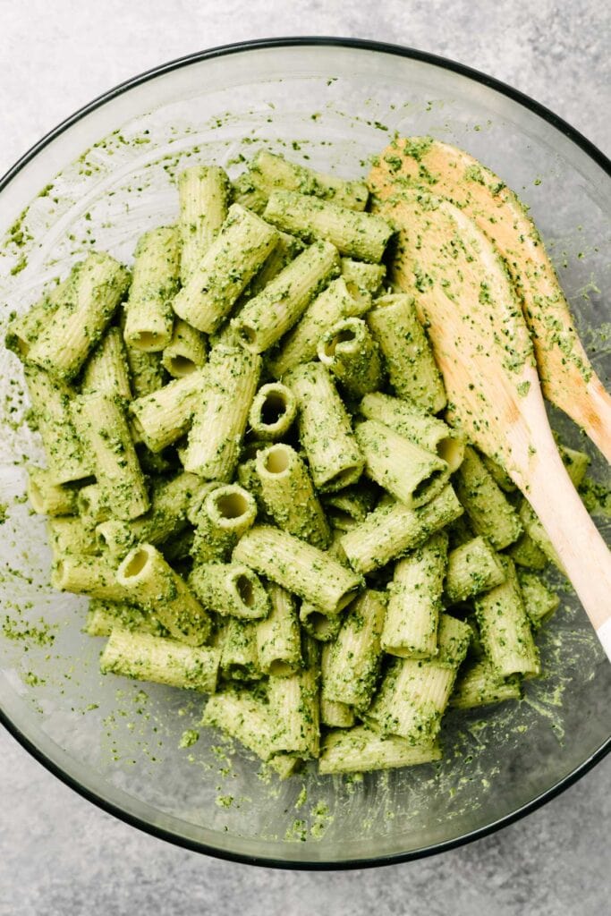 Cooked rigatoni pasta tossed with spinach pesto in a large glass mixing bowl.