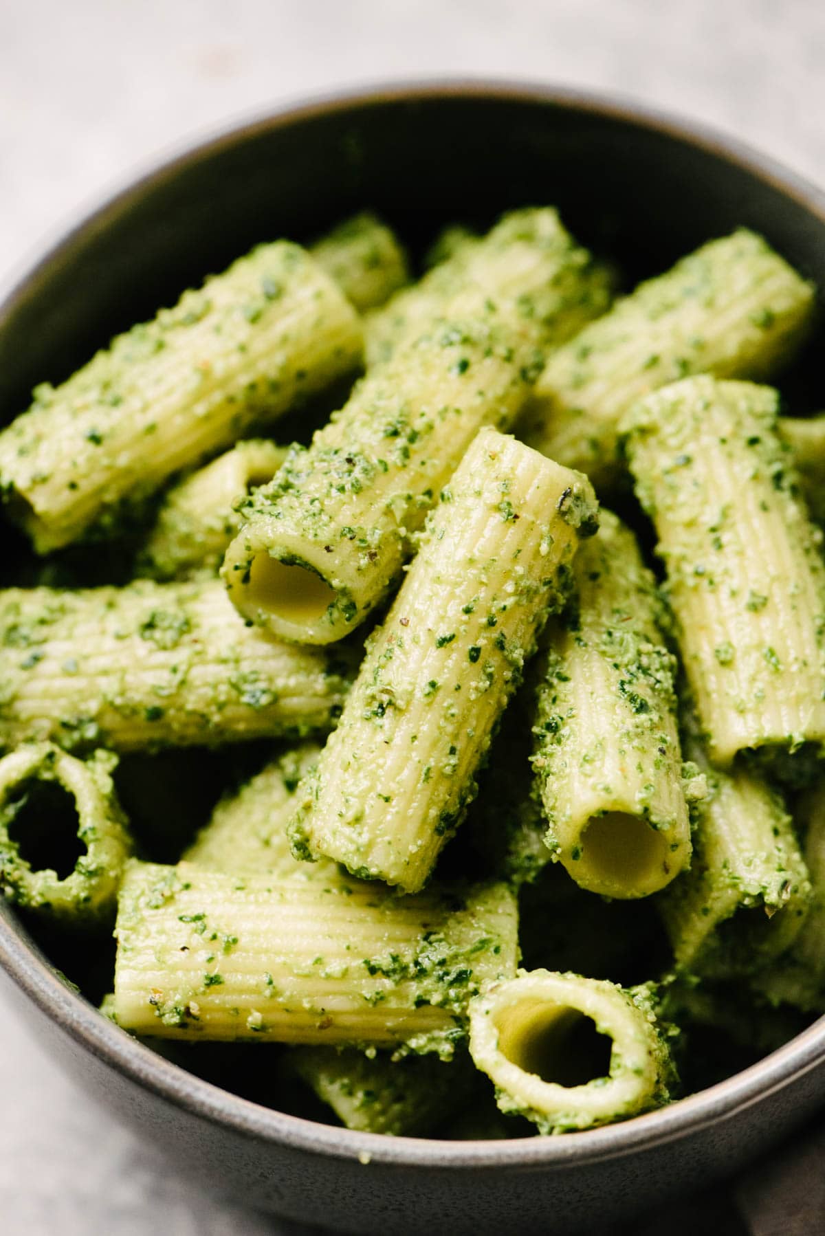Side view, cooked pasta tossed with creamy pesto sauce in a dark grey bowl.