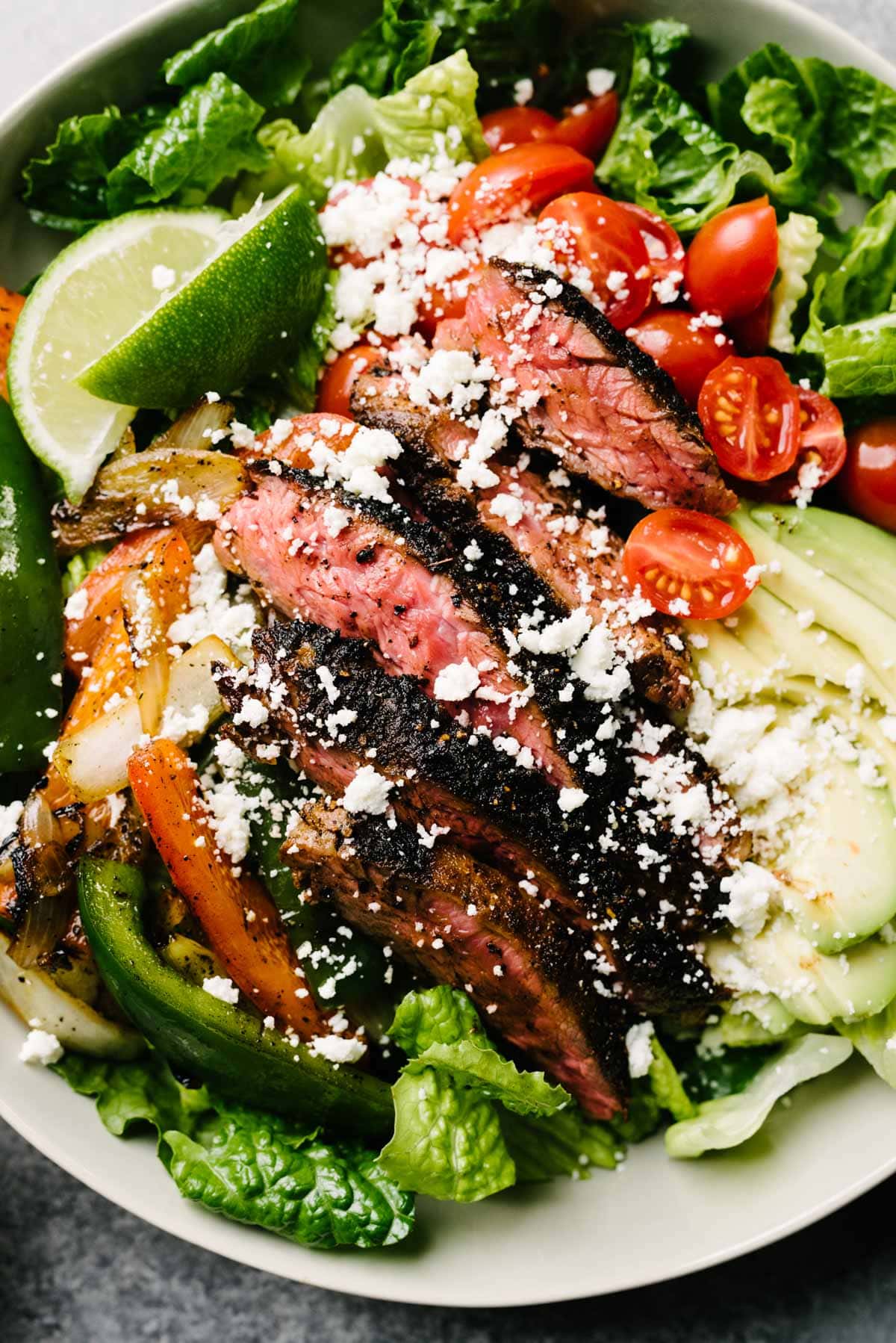 A close-up image of steak fajita salad with seared steak and fajita veggies over romaine lettuce, bell peppers, and sliced avocado, garnished with crumbed queso fresco.