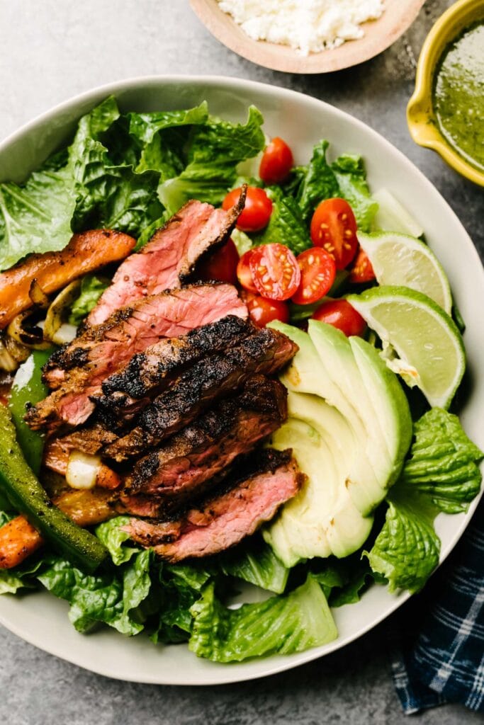 Composed fajita salad with steak in a low tan bowl - chopped romaine, cherry tomatoes, avocado, sliced steak, and sauteed vegetables - with queso fresco cheese and cilantro lime vinaigrette on the side.