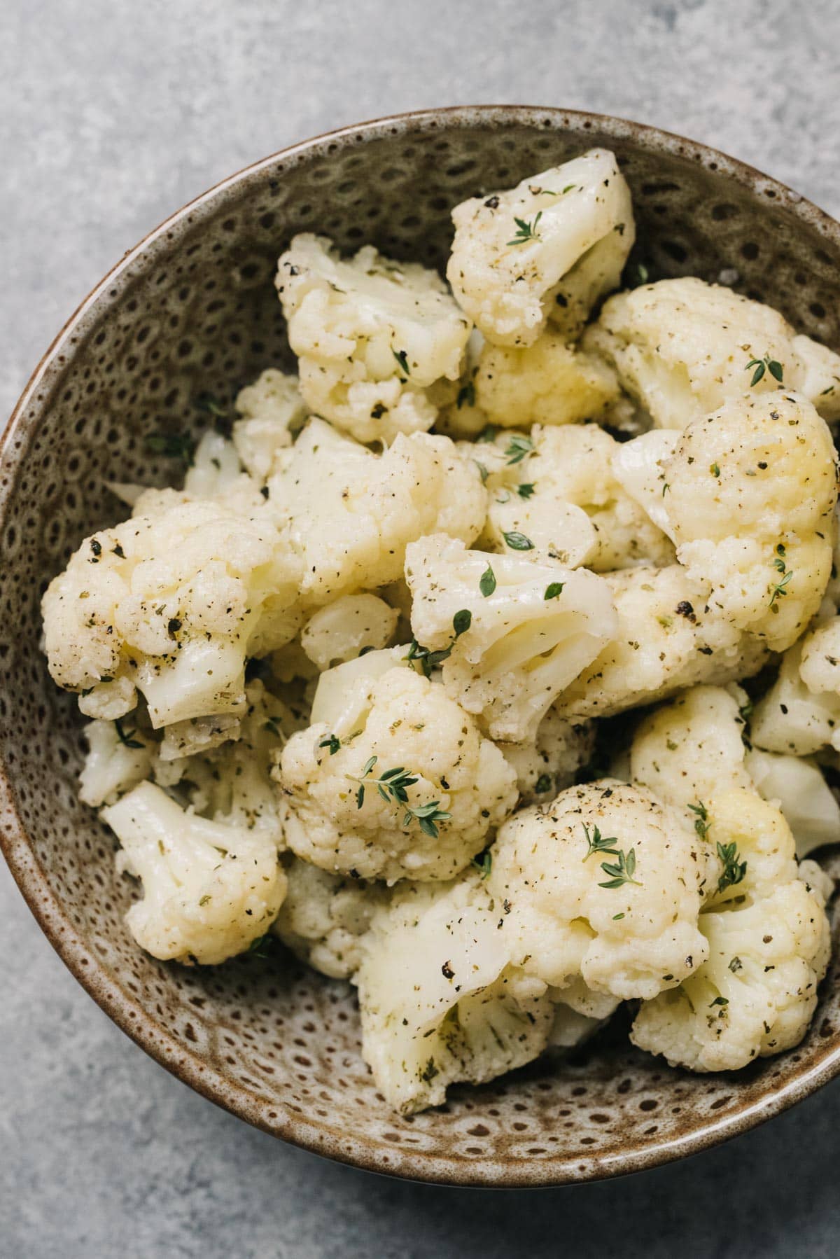 Steamed cauliflower florets tossed with olive oil, Italian seasoning, and fresh thyme in a brown speckled bowl on a concrete background.