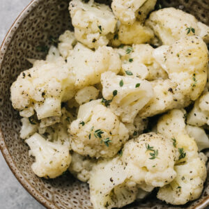Steamed cauliflower florets tossed with olive oil, Italian seasoning, and fresh thyme in a brown speckled bowl on a concrete background.