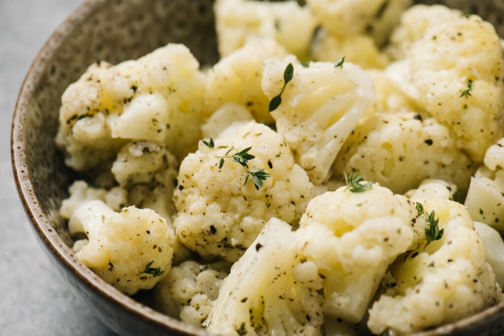 Side view, steamed cauliflower tossed with olive oil, Italian seasoning, and fresh thyme in a brown speckled bowl.
