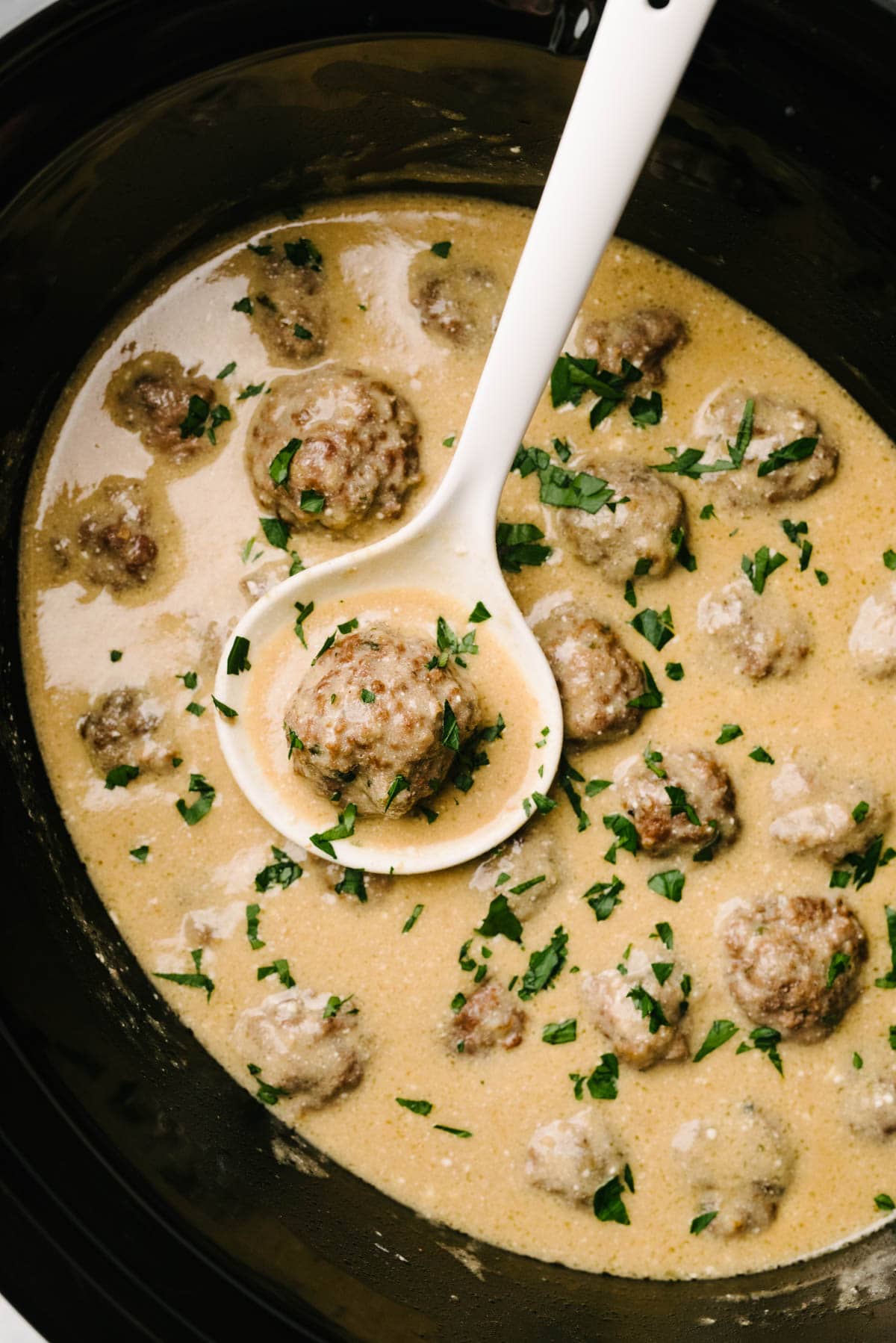 A white ladle tucked into a crockpot of Swedish meatballs in a creamy sauce, garnished with fresh parsley.