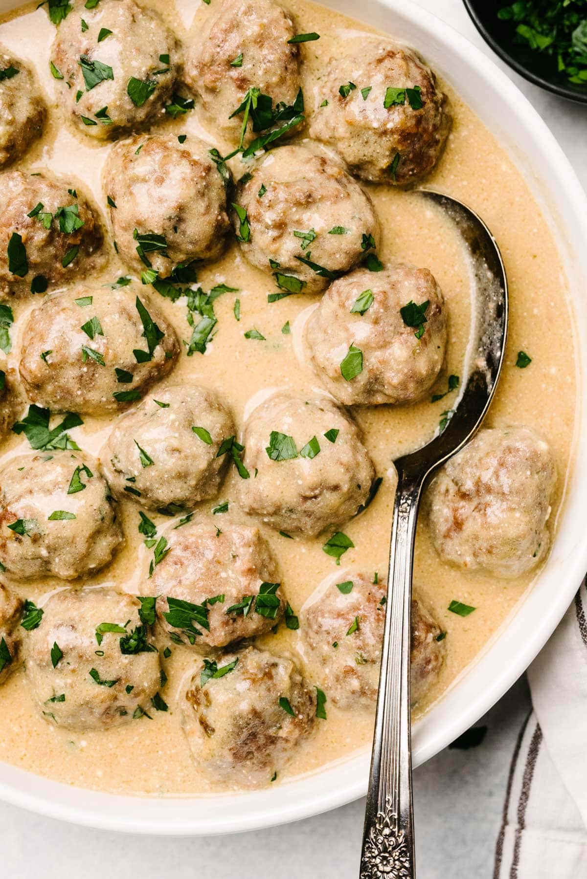 A spoon tucked into a serving bowl filled with Swedish meatballs and a creamy sauce.