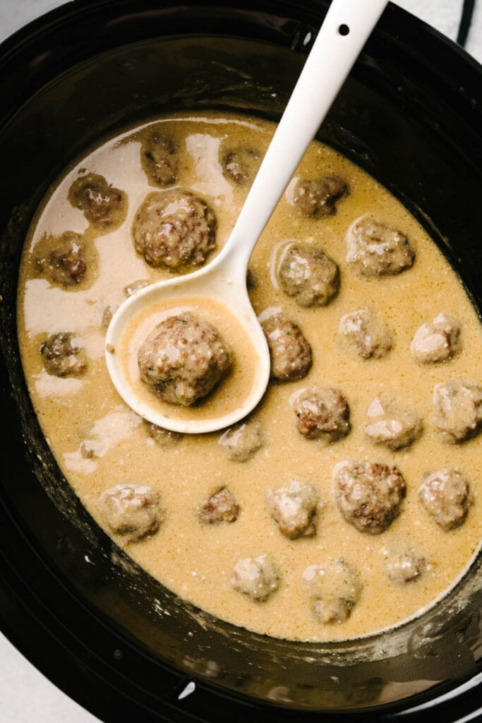 A white ladle tucked into a crockpot of Swedish meatballs in a creamy sauce.