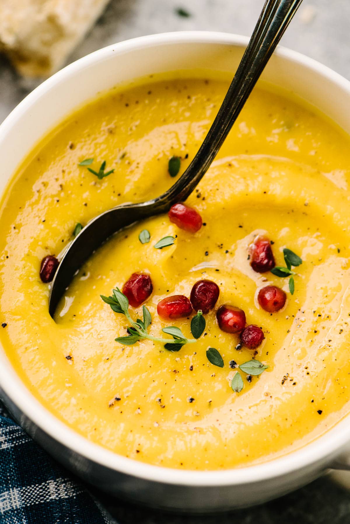 Side view, a soup spoon tucked into a bowl of roasted butternut squash soup garnished with pomegranate seeds and fresh thyme leaves.