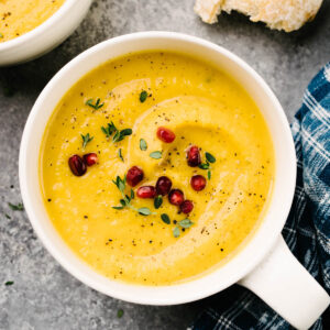 Two bowls of roasted butternut squash soup garnished with pomegranate seeds and fresh thyme on a cement background, with hunks of bread and a blue patterned napkin to the side.