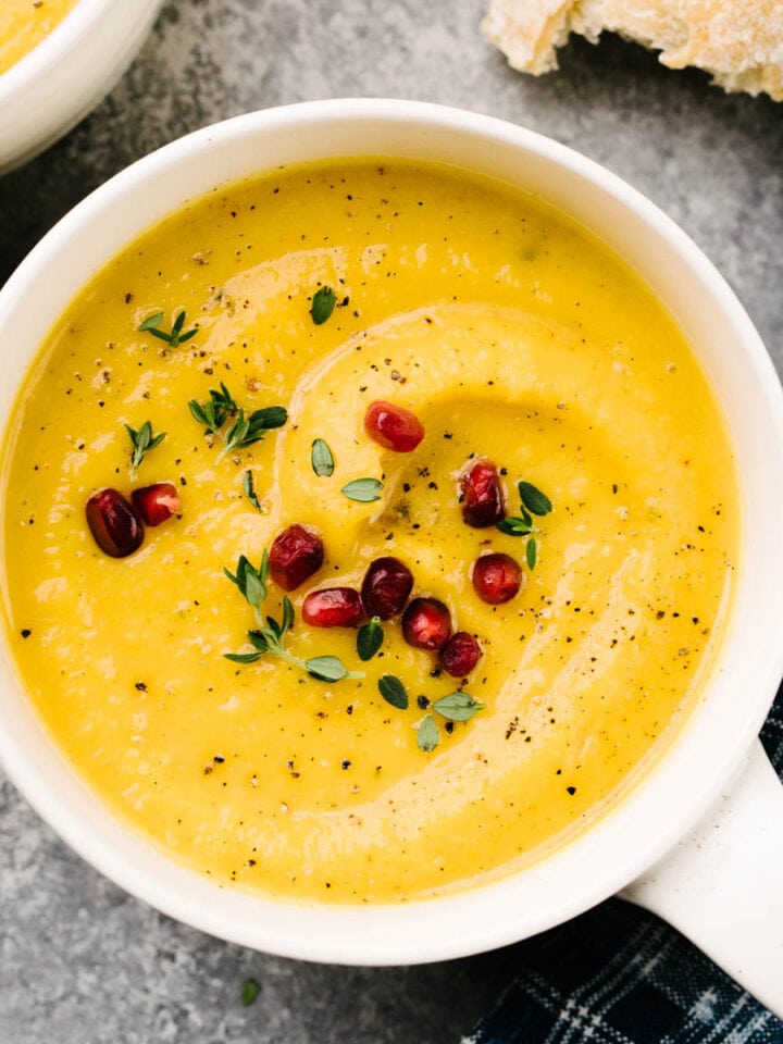 Two bowls of roasted butternut squash soup garnished with pomegranate seeds and fresh thyme on a cement background, with hunks of bread and a blue patterned napkin to the side.