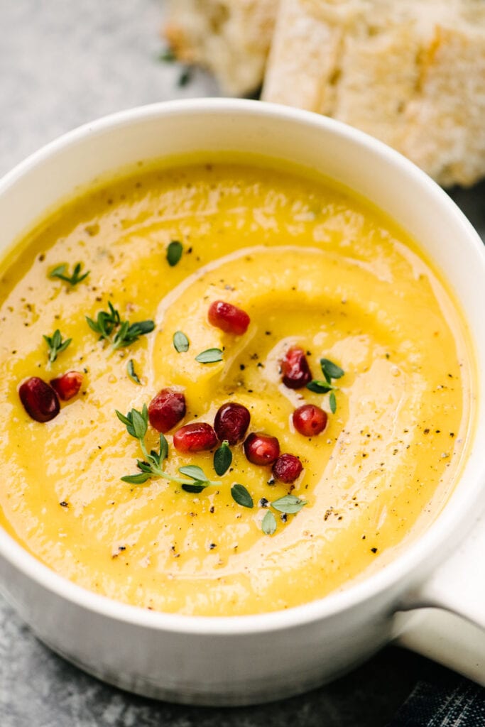 Side view, roasted butternut squash soup in a white bowl, garnished with fresh thyme leaves and pomegranate seeds.