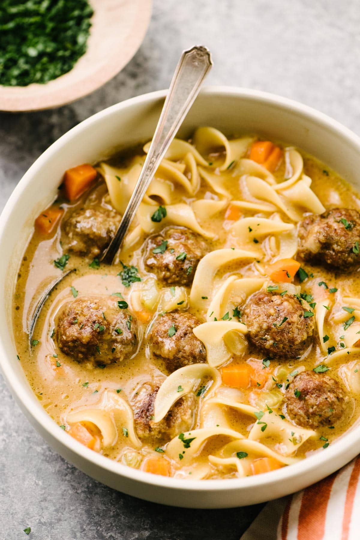 Side view, a bowl of Swedish meatball soup on a cement background with a pinch bowl of chopped fresh parsley in the background.