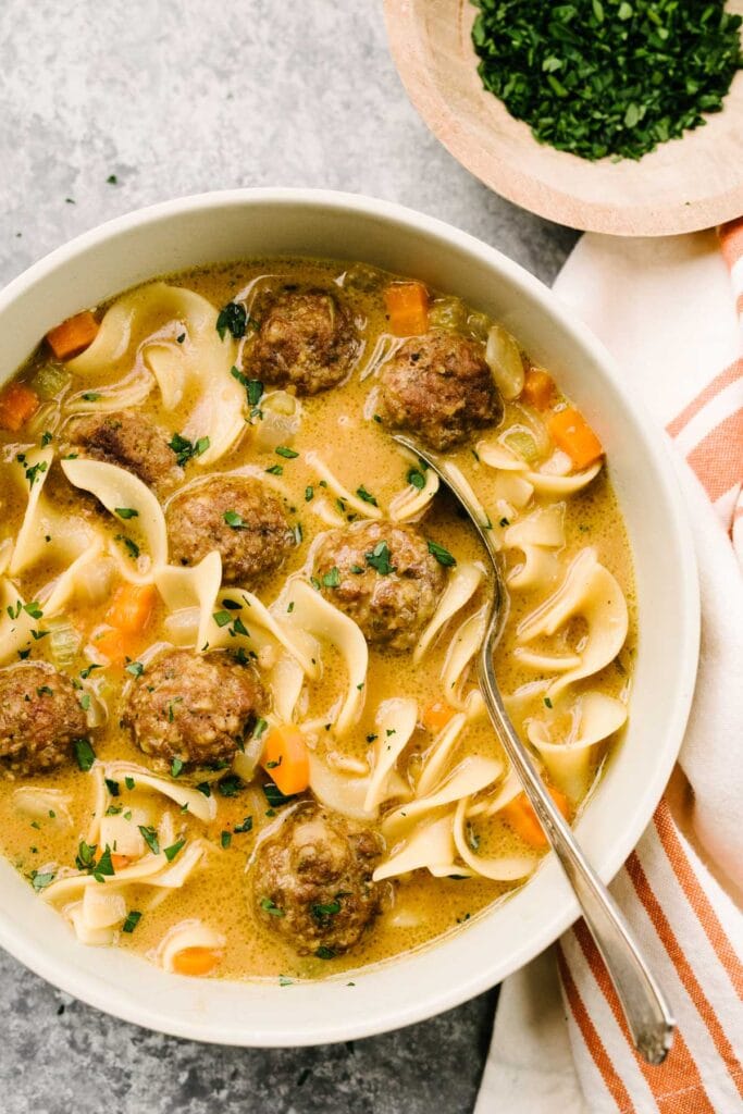 A spoon tucked into a bowl of Swedish meatball soup on a cement background, with a striped linen napkin and bowl of fresh chopped parsley to the side.