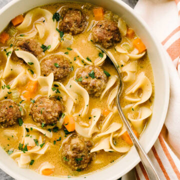 A spoon tucked into a bowl of Swedish meatball soup, garnished with fresh parsley, with a striped linen napkin to the side.