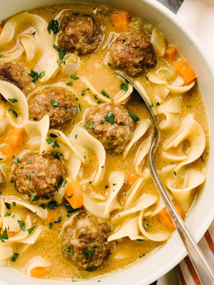 A spoon tucked into a bowl of Swedish meatball soup, garnished with fresh parsley, with a striped linen napkin to the side.