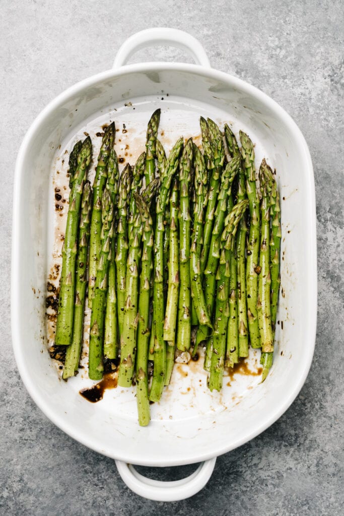 Trimmed asparagus spears tossed with balsamic vinegar, olive oil, salt, and pepper in a white casserole dish.