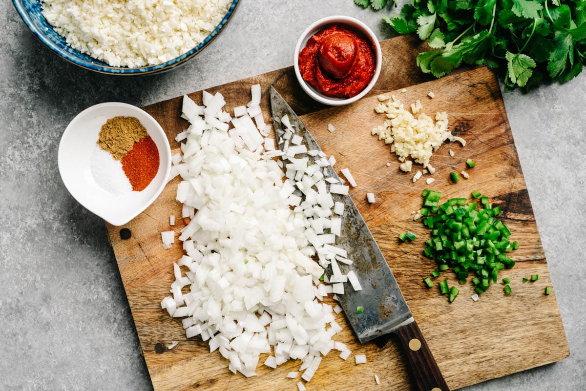The ingredients for Mexican cauliflower rice arranged across a cutting board - cauliflower rice, white onion, garlic, jalapeno, tomato paste, cilantro, and seasonings.
