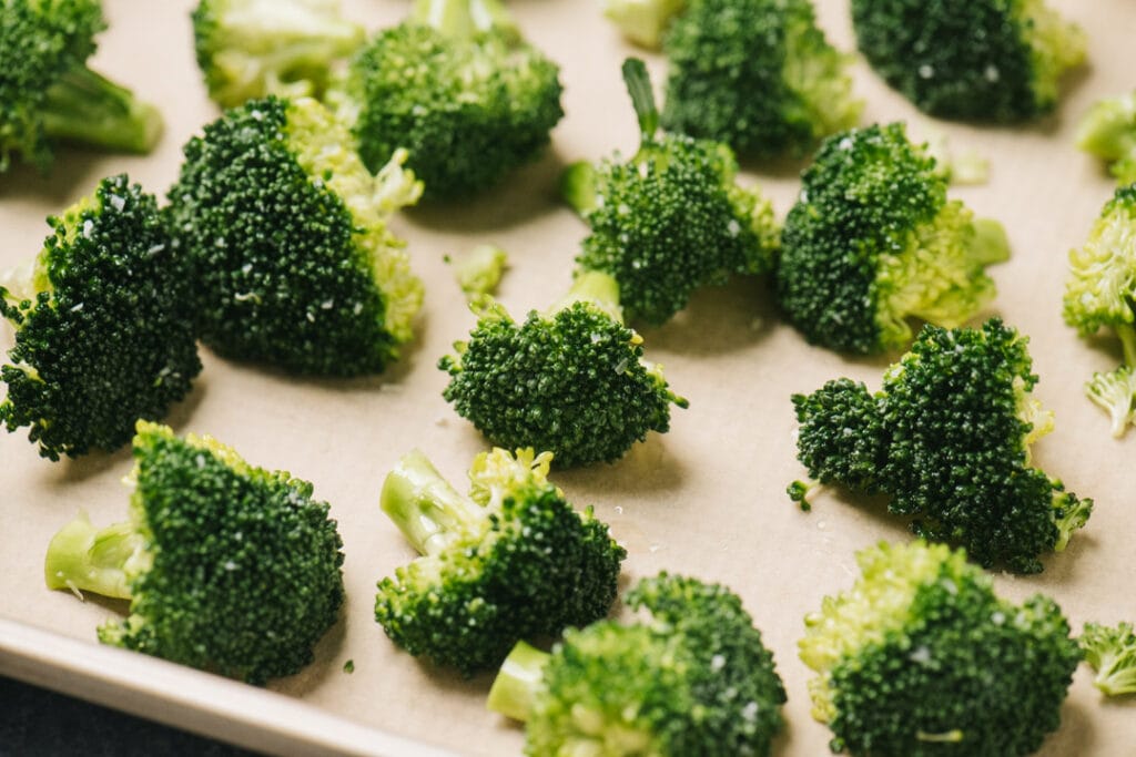 Side view, broccoli florets evenly spaced on a parchment lined baking sheet before going into the oven.