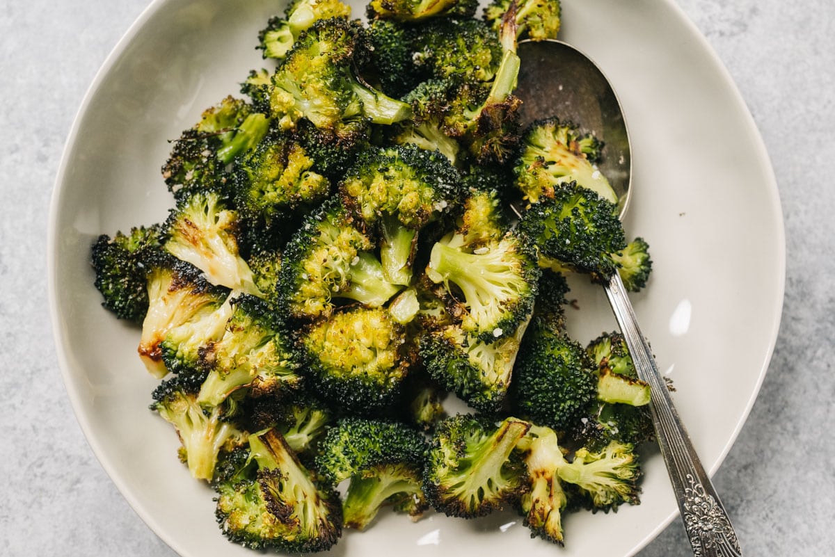 Oven roasted broccoli florets in a low tan bowl with a vintage silver serving spoon on a concrete background.