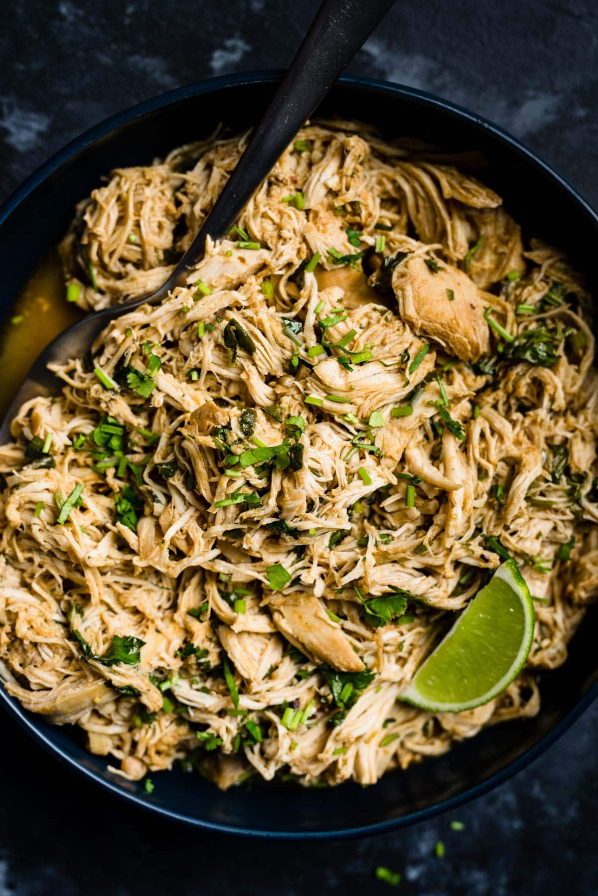 A serving spoon tucked into a bowl of shredded slow cooker cilantro lime chicken, garnished with a lime wedge and lots of fresh cilantro.