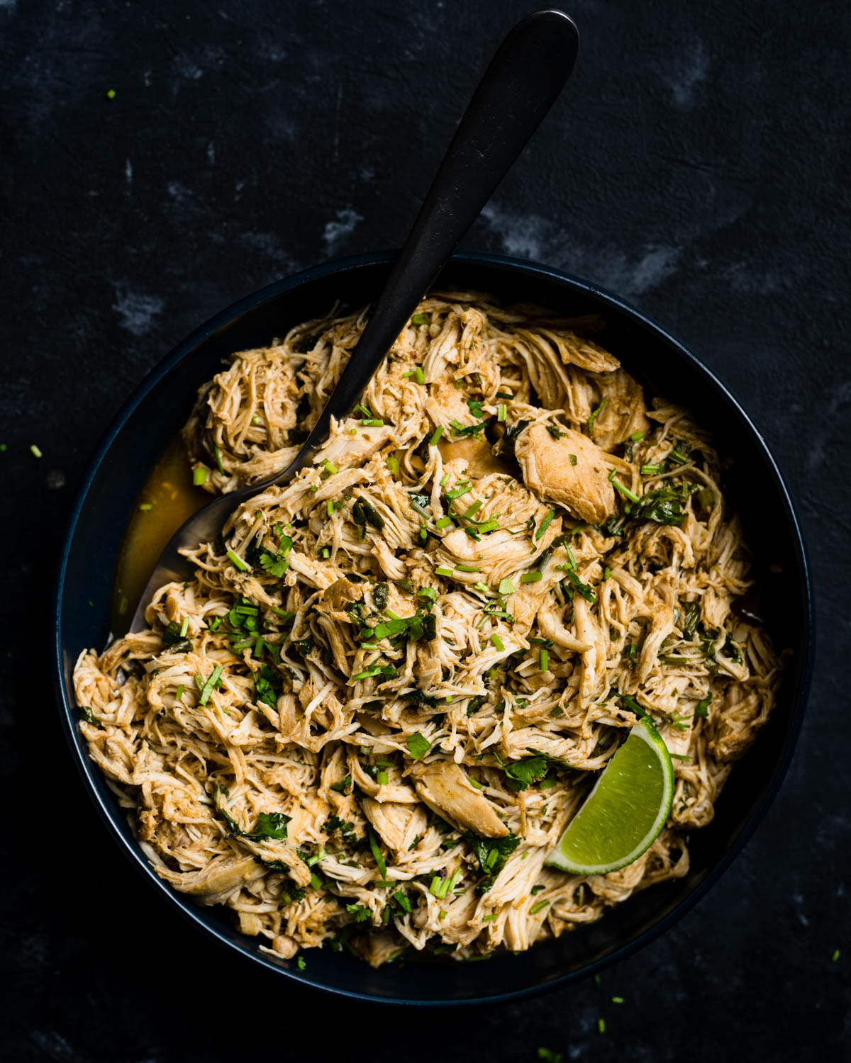 A bowl of shredded slow cooker cilantro lime chicken on a dark black chalkboard background.