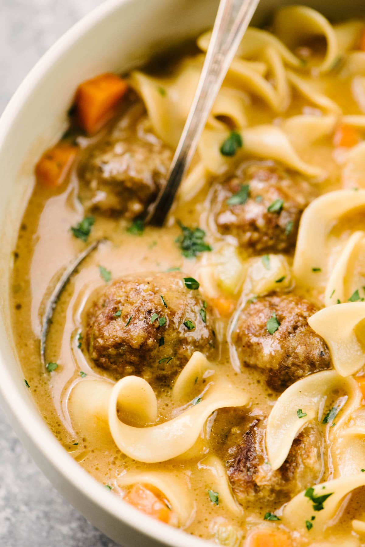 Side view, a spoon tucked into a bowl of Swedish meatball soup with egg noodles, garnished with fresh parsley.