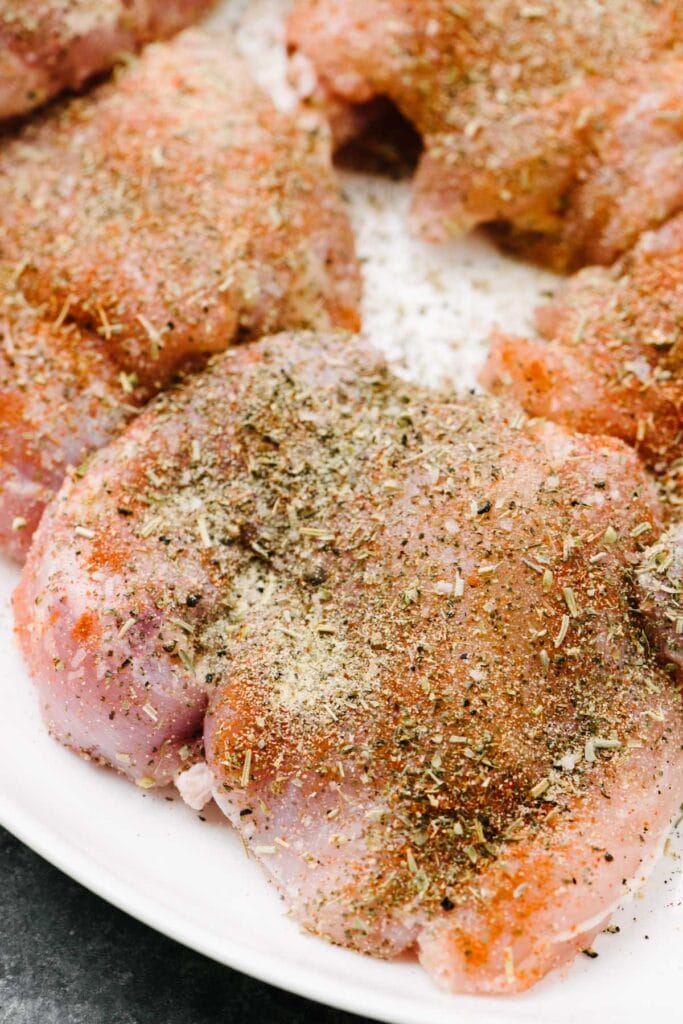 Side view, raw thicken thighs on a white plate seasoned with garlic powder, Italian seasoning, salt, pepper, and paprika.