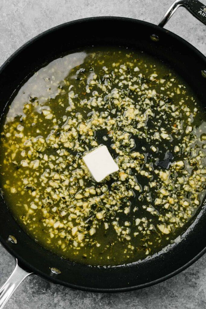 Adding butter to a skillet with garlic, thyme, and chicken broth to create a pan sauce.
