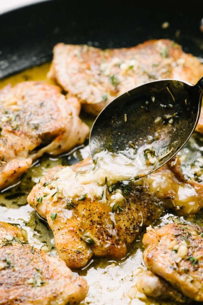 Side view, spooning garlic butter sauce over seared chicken thighs in a skillet.