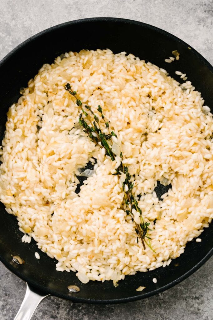 Partially cooked risotto with a thyme sprig in a skillet.